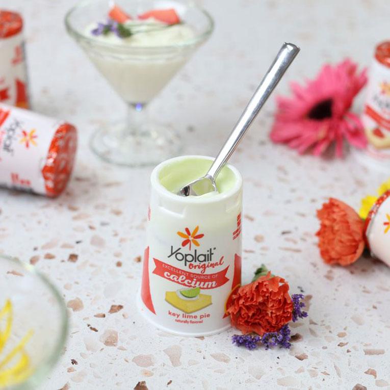 An open tub of Yoplait Original Key Lime Pie flavor with a teaspoon in it sitting on a table with a drink, yogurt tubs and flowers also on the table.