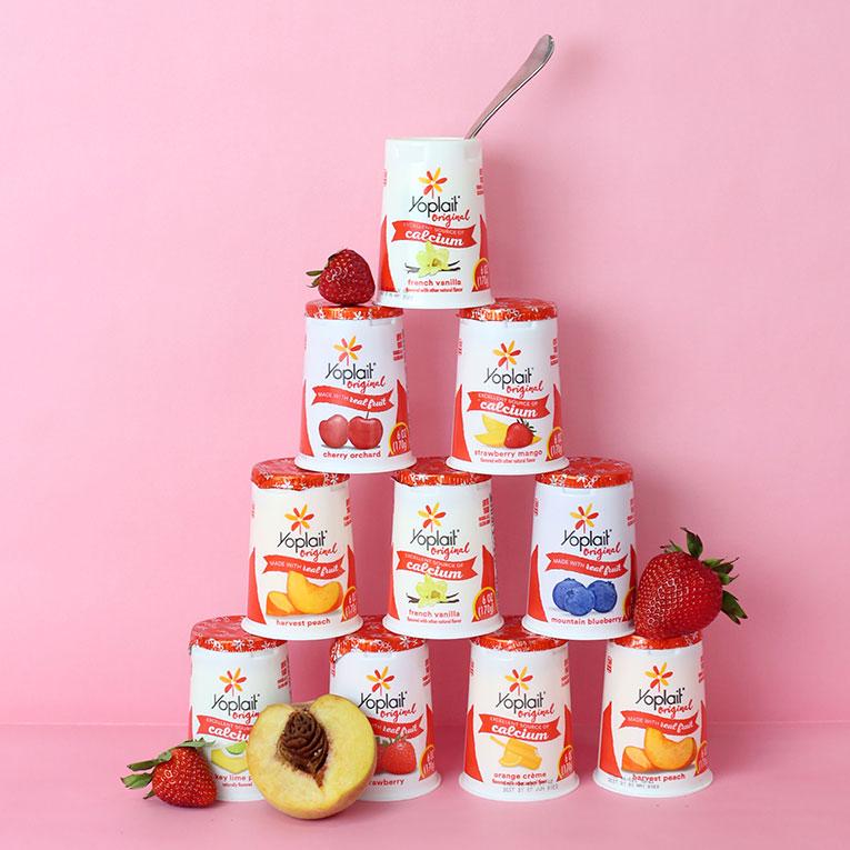 10 tubs of Yoplait Original yogurt flavors arranged in a pyramid with a teaspoon in the top tub and fruit around the pyramid.