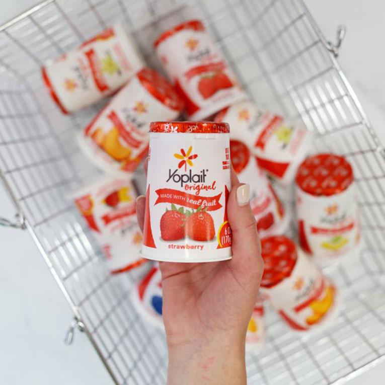 A hand holding a tub of Yoplait Original Strawberry flavor over a shopping basket full of Yoplait Original yogurt tubs.