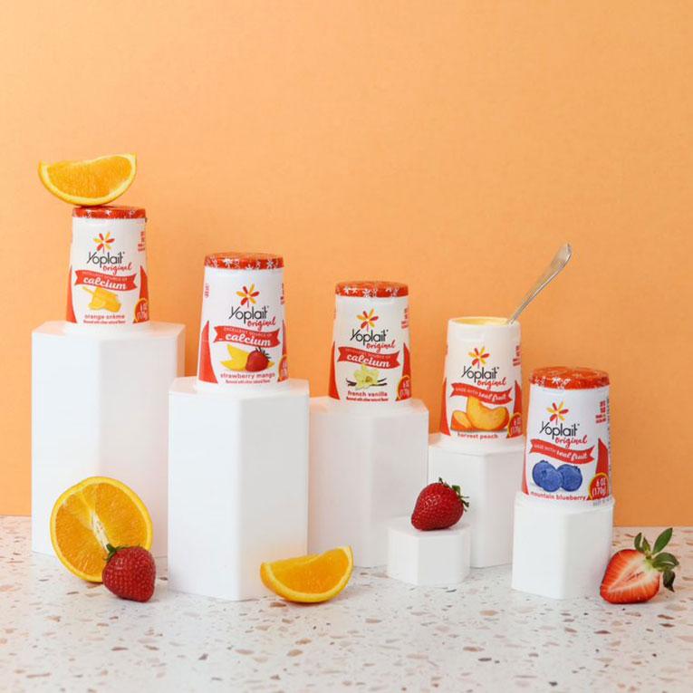 Five tubs of Yoplait Original yogurt arranged on white stands of different sizes on a table, surrounded by fruit.